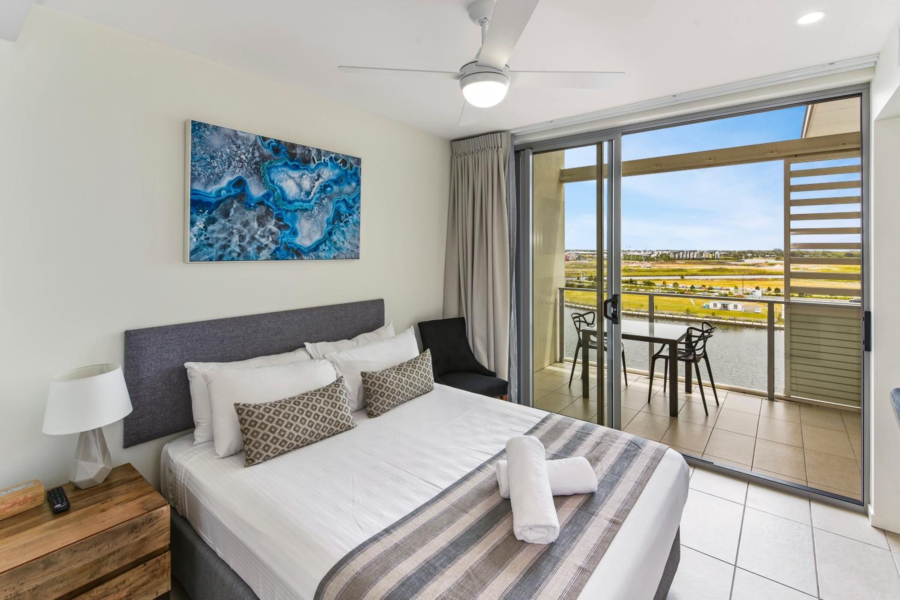 A hotel room with a bed, a painting, a ceiling fan, a nightstand, a lamp, and a balcony with a view of the city.