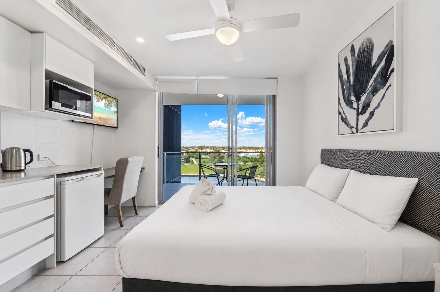 A modern hotel room with a bed, a kitchenette, a dining area, and a balcony with a view.