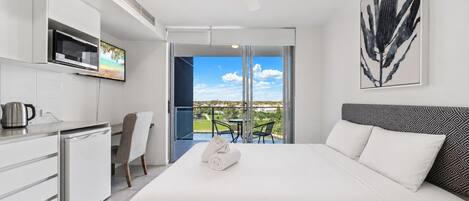 A modern hotel room with a bed, a kitchenette, a dining area, and a balcony with a view.