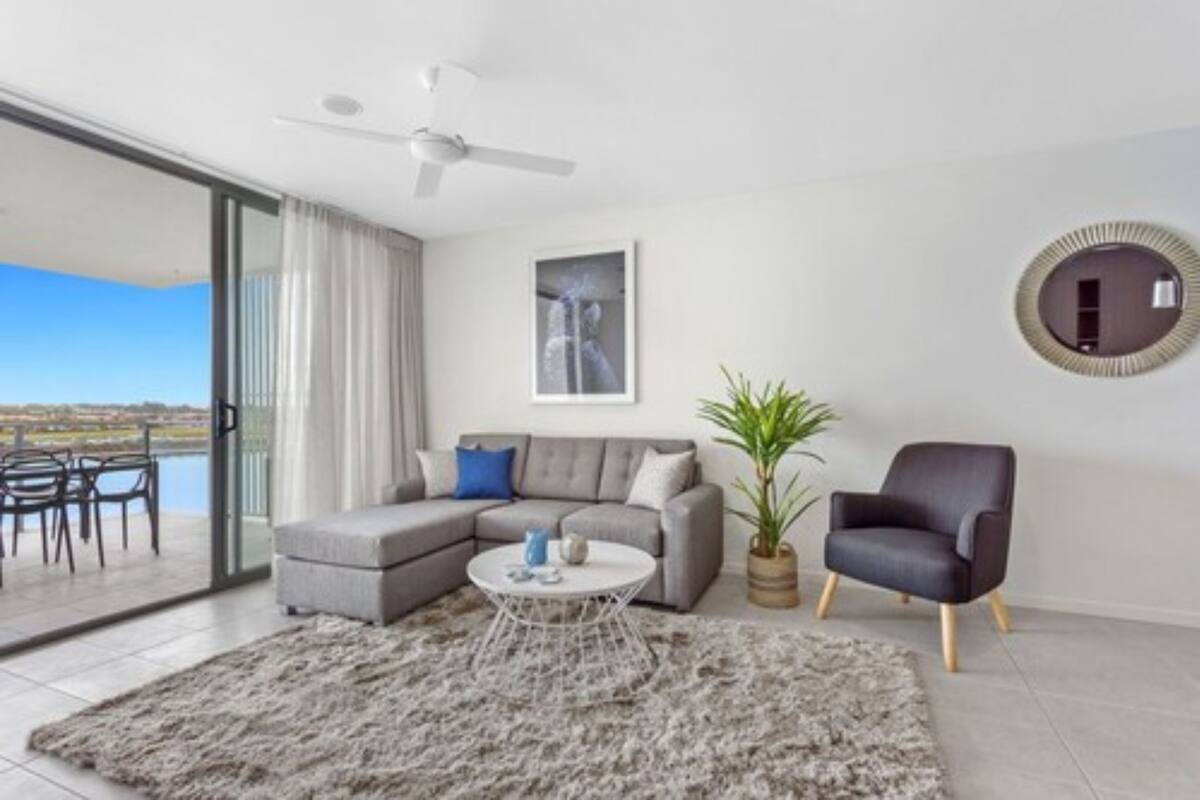 Een moderne woonkamer met een grijze hoekbank, een ronde salontafel, een potplant, een zwarte fauteuil en een plafondventilator.