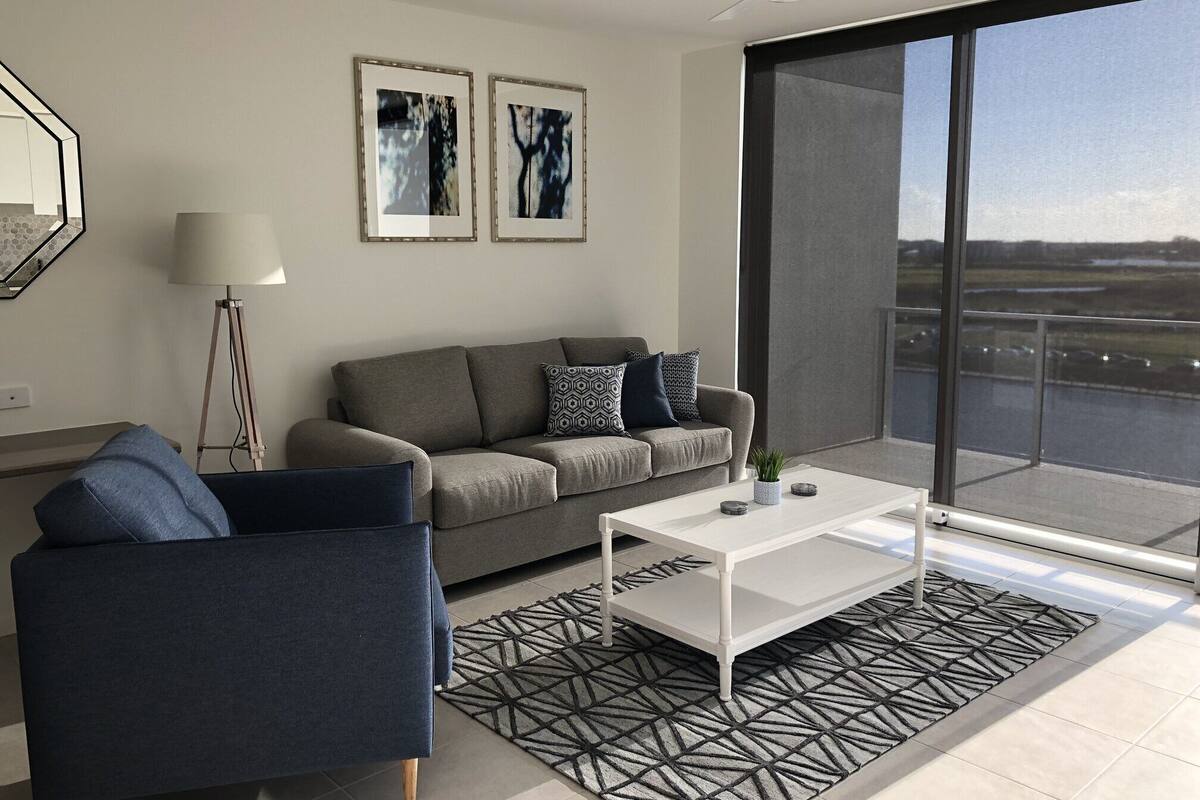 Een moderne woonkamer met een grijze bank, een blauwe fauteuil, een witte salontafel en een vloerkleed met dessin. Twee ingelijste abstracte schilderijen hangen aan de muur, er staat een vloerlamp en een groot raam biedt uitzicht op een waterpartij en gebouwen.