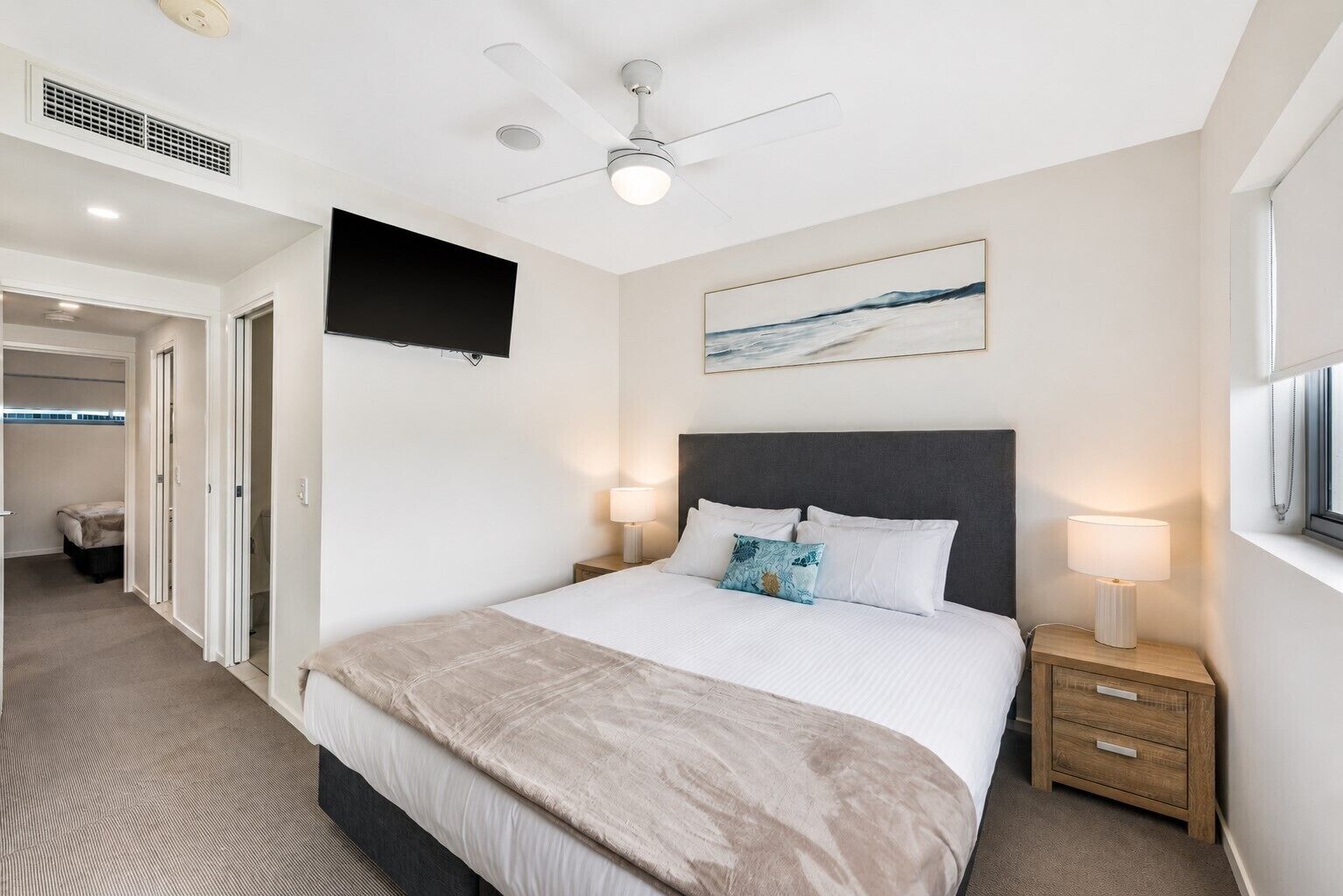 A hotel room with a bed, bedside lamps, a ceiling fan, a mounted TV, and a painting on the wall.