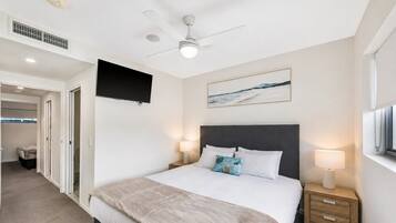 A hotel room with a bed, bedside lamps, a ceiling fan, a mounted TV, and a painting on the wall.