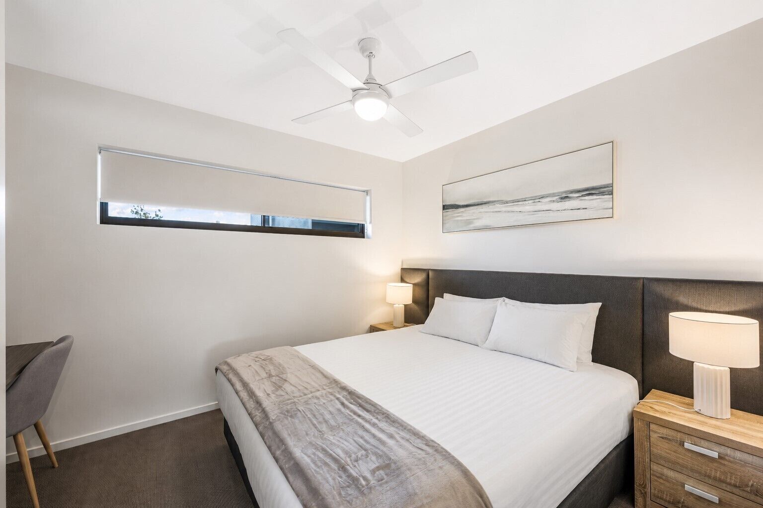A hotel room with a bed, bedside lamps, a ceiling fan, a window with blinds, and a painting on the wall.