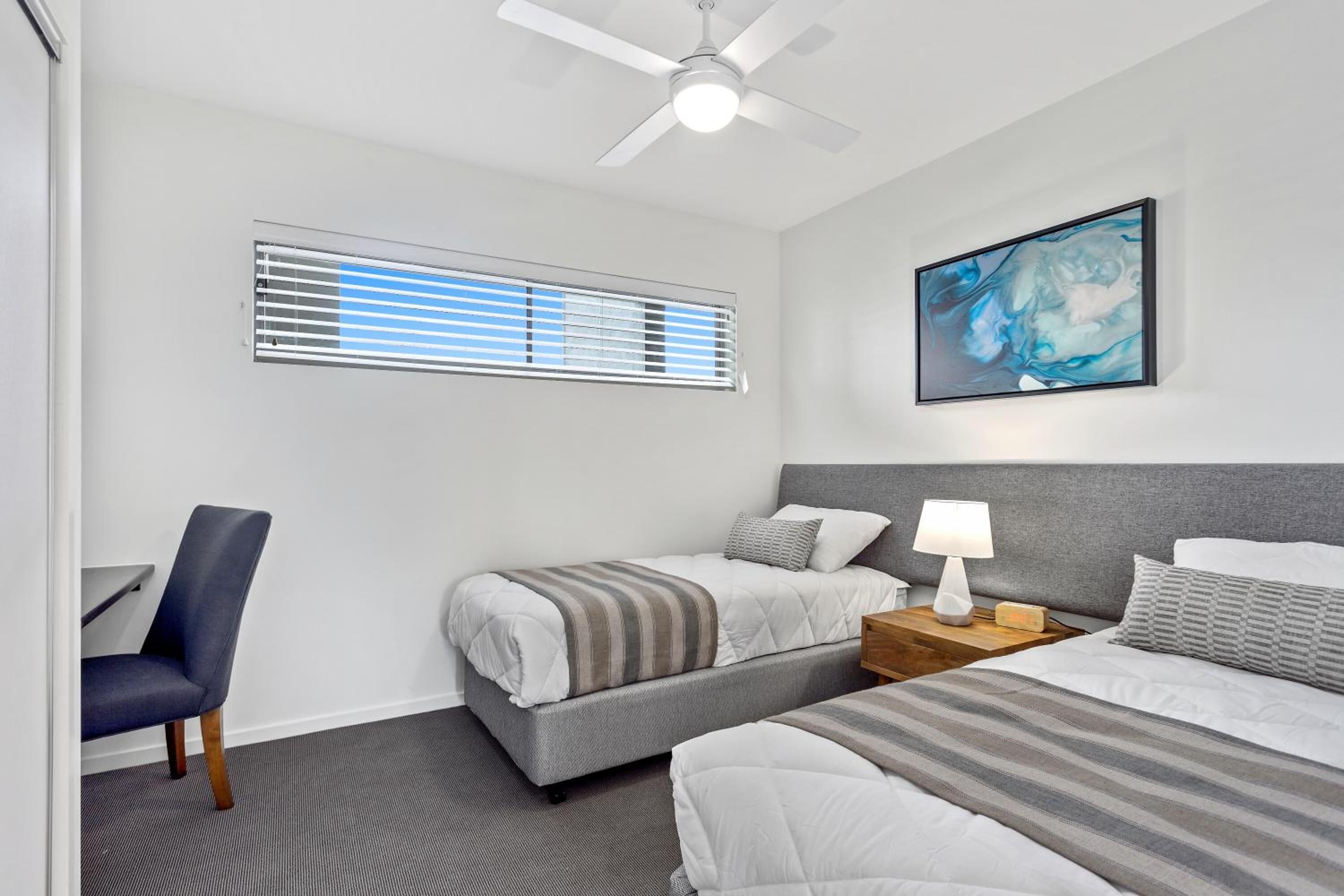 A hotel room with two beds, a desk, a chair, a ceiling fan, a window with blinds, and a painting on the wall.