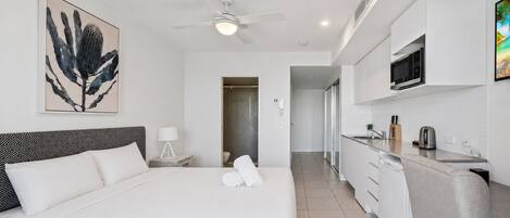 A modern hotel room with a bed, a ceiling fan, a bathroom, and a kitchenette.
