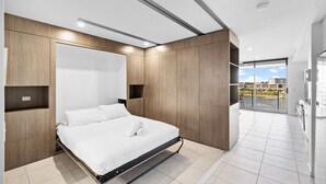 A modern bedroom with a bed, wooden paneling, and a view of a cityscape through a large window. - North Shore Kawana and Waterline (Birtinya)