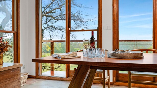 Un comedor con una mesa de madera, cristalería transparente y una botella, con vistas a árboles y cielo a través de amplios ventanales.