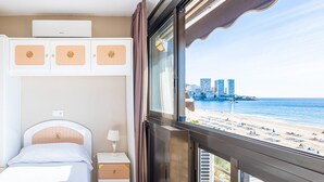 Une chambre d’hôtel avec un lit, une commode, une table de chevet et une fenêtre donnant sur la plage et le paysage urbain.