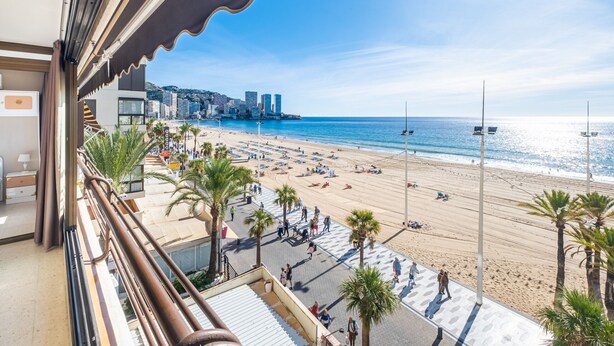 Une vue sur la plage depuis un balcon, avec des palmiers, une promenade et un paysage urbain en arrière-plan.