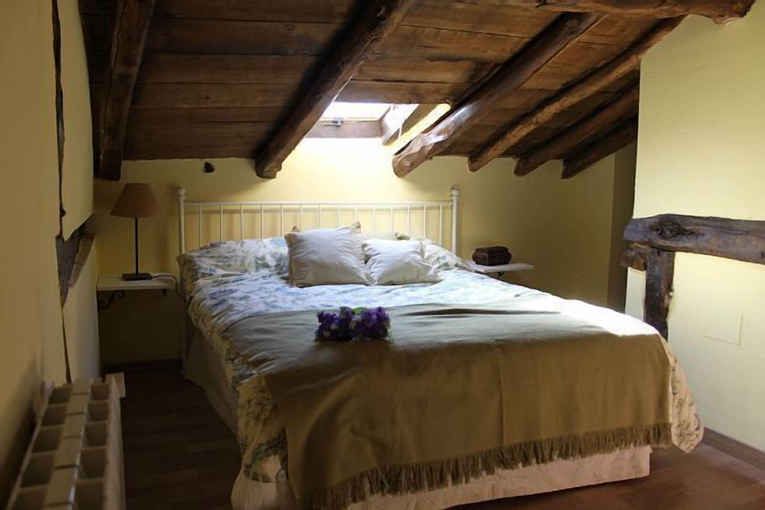 Ein Schlafzimmer mit Holzbalkendecke, einem Bett mit weißer Bettwäsche, einem Nachttisch mit Lampe und einem kleinen Tisch mit einer Schüssel.