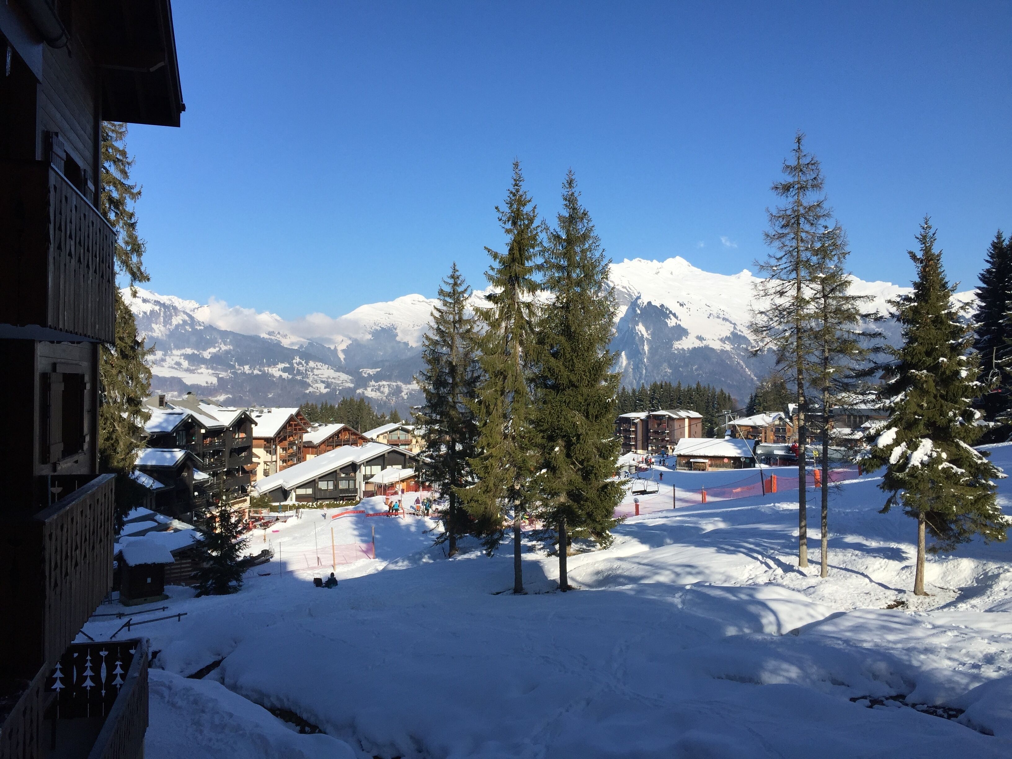 A snow-covered mountain village with wooden buildings and snow-covered roofs, surrounded by tall evergreen trees and a clear blue sky.