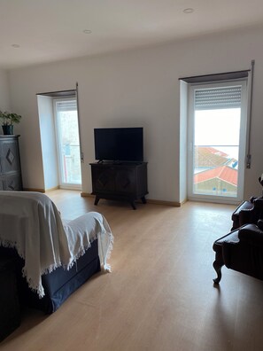 Et rummeligt værelse med et stort vindue, et fladskærms-tv på et træskab, en seng med et hvidt lagen og en mørk sofa.