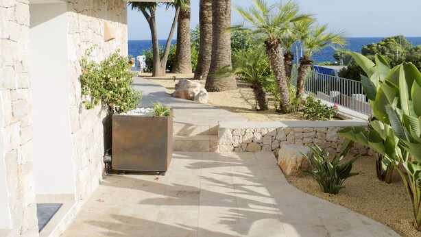 A pathway leading to a white building with stone walls, palm trees, and a view of the sea. - Wonderful luxury villa in 1st line of the sea in Moraira with private pool. (Teulada / Moraira)