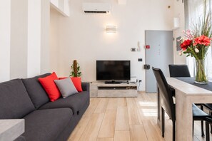 A modern living room with a flat-screen TV, a grey sofa with red and grey cushions, a dining table with chairs, and a vase with flowers. - I FRAGOLI A SANTA CROCE (Firenze)