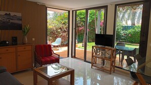 A living room with a glass coffee table, a red sofa, a TV on a wooden stand, and a view of a garden through large sliding doors.