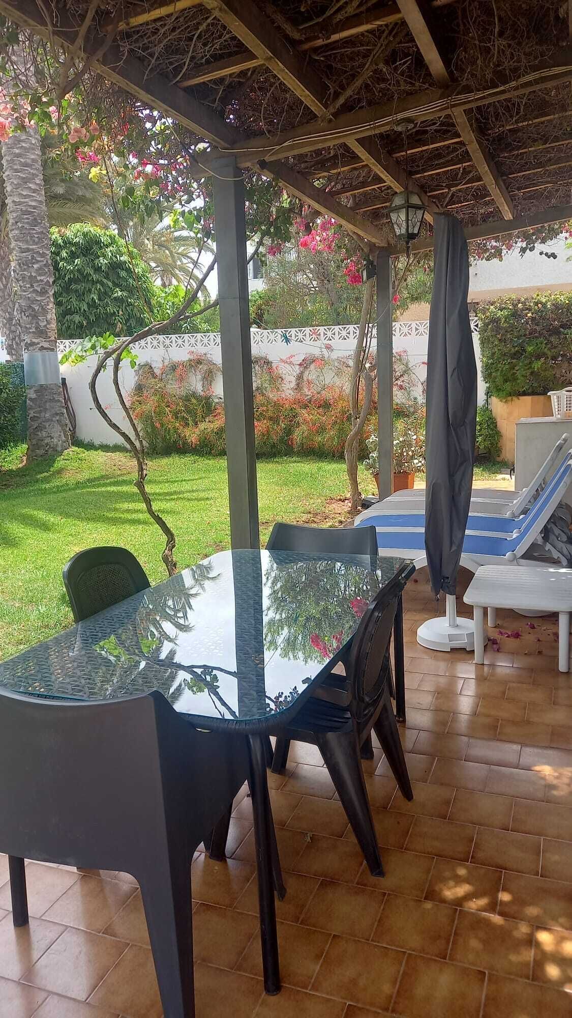 Un patio al aire libre con una mesa de vidrio, sillas negras y una zona junto a la piscina con sillones de descanso y una sombrilla.