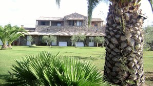 A stone building with a tiled roof, surrounded by a well-maintained lawn and palm trees. - DomusAntiga three-room apartment 10 minutes from the most beautiful beaches of the coast!Wifi (San Giovanni Suergiu)
