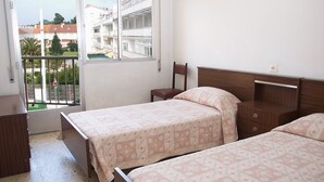 A bedroom with two beds, a wooden nightstand, a chair, and a window with a view of buildings and greenery.