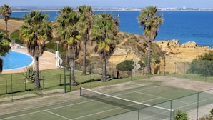 A tennis court with a net, surrounded by palm trees and a pool, overlooking a beach and ocean. - Magnificently located apartment with sea view (Lagos)