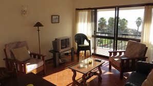 A living room with a sofa, armchairs, a coffee table, a television, and a balcony with a view of palm trees. - Magnificently located apartment with sea view (Lagos)