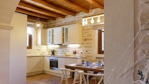 Une cuisine avec des armoires blanches, un plafond en bois et un mur en pierre. On y trouve une table avec des chaises et un luminaire suspendu.