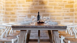 Une table à manger, agrémentée de verres à vin et d’une bouteille, est entourée de chaises blanches dont les assises sont en bois.