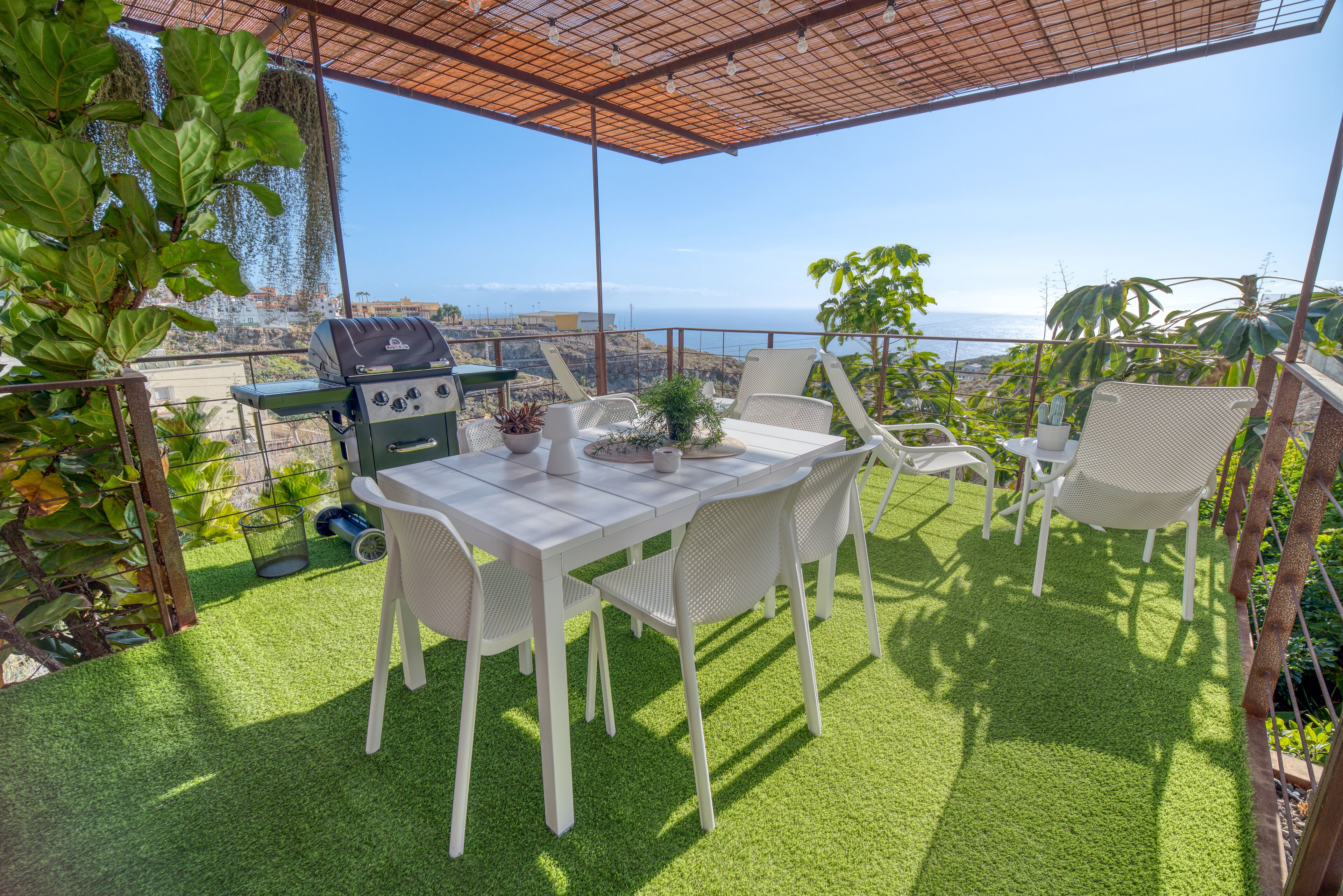 An outdoor patio with a grill, white chairs, and a table, overlooking a scenic view.