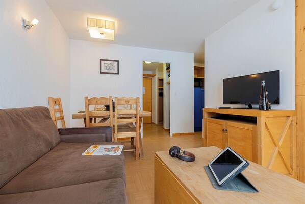 A living room with a brown sofa, wooden chairs, a dining table, a television, and a small table with a tablet and headphones.