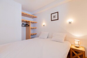 A bedroom with a bed, a wooden nightstand with a lamp, a wall-mounted shelf with clothes, and a framed picture.