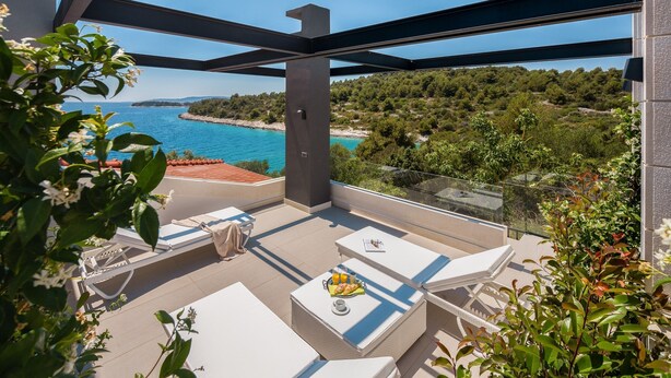 A balcony with white lounge chairs, a table with a fruit basket, and a view of the sea and greenery. - Villa Argemonia – Luxury Beachfront Escape in Tatinje Bay (Okrug Gornji)