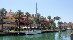A marina with a sailboat docked, surrounded by multi-story buildings with balconies and palm trees.