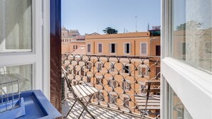 Un balcón con barandilla de metal, vistas a edificios cercanos y cielo despejado.