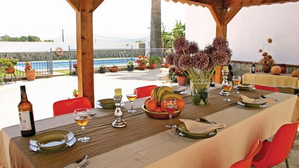 An outdoor dining area with a table set for two, featuring a bottle of wine, glasses, a bowl of fruit, and a vase of flowers.