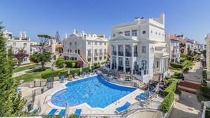 Hotel met zwembad, omringd door ligbedden en parasols, een aangelegde tuin en helderblauwe lucht.