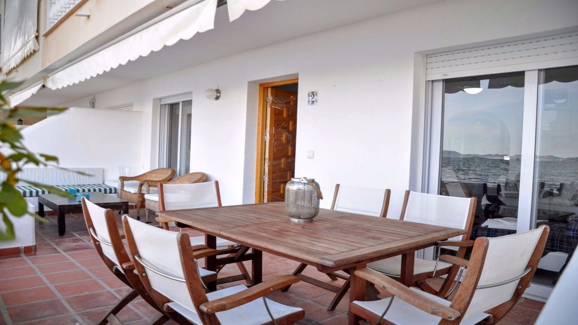 A wooden table with eight chairs, a large vase, and a view of the sea through a glass door.