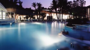 A swimming pool with lounge chairs and palm trees, surrounded by buildings with warm lighting. - Beautiful house in La Manga The pedruchillo. First line beach garage and pool (San Javier)