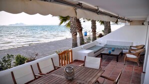 A terrace with a view of the sea, featuring a wooden table, white cushioned seating, and a wicker chair. - Beautiful house in La Manga The pedruchillo. First line beach garage and pool (San Javier)