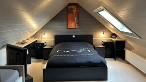 Een moderne slaapkamer met een schuine kap, een bed met een zwart hoofdeinde, twee nachtkastjes met lampen, een poster aan de muur en een dakraam.