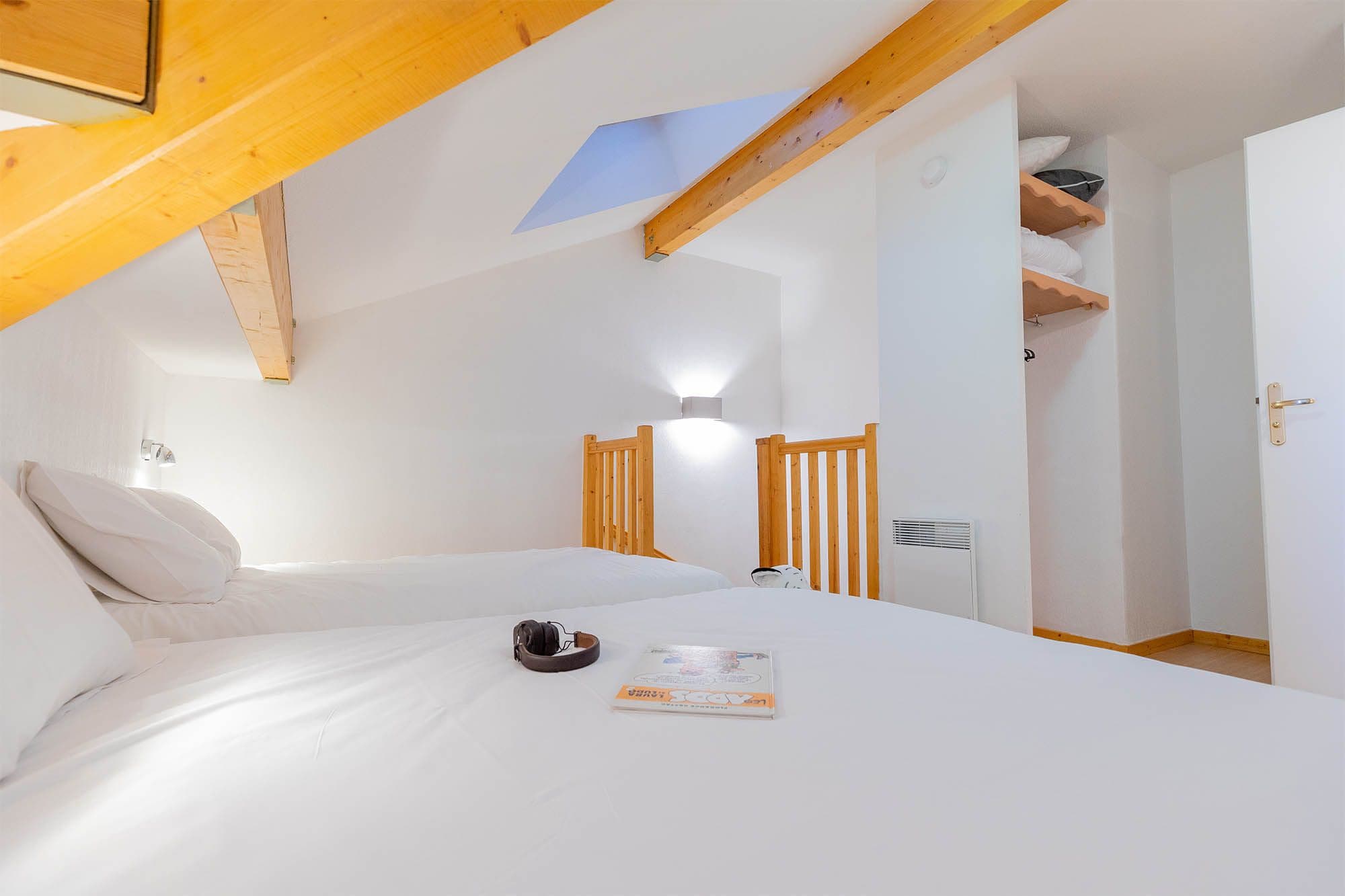 A bedroom with a sloped ceiling, a bed with white bedding, a wooden headboard, a nightstand, a wall-mounted lamp, and a door with a towel rack.