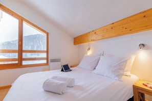 A bedroom with a large window, a bed with white linens, a bedside table with a lamp, and a wall-mounted heater.