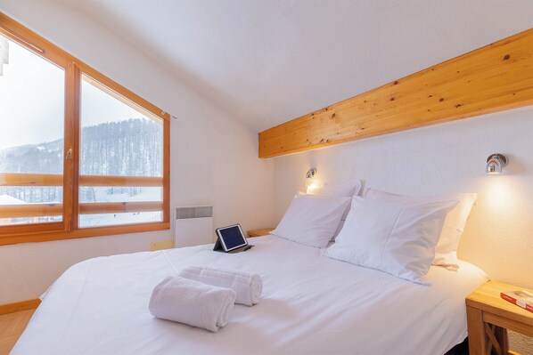 A bedroom with a large window, a bed with white linens, a bedside table with a lamp, and a wall-mounted heater.