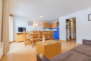 A living room with a sofa, a dining area with chairs and a table, a kitchen with cabinets and a TV, and a hallway leading to a bedroom.