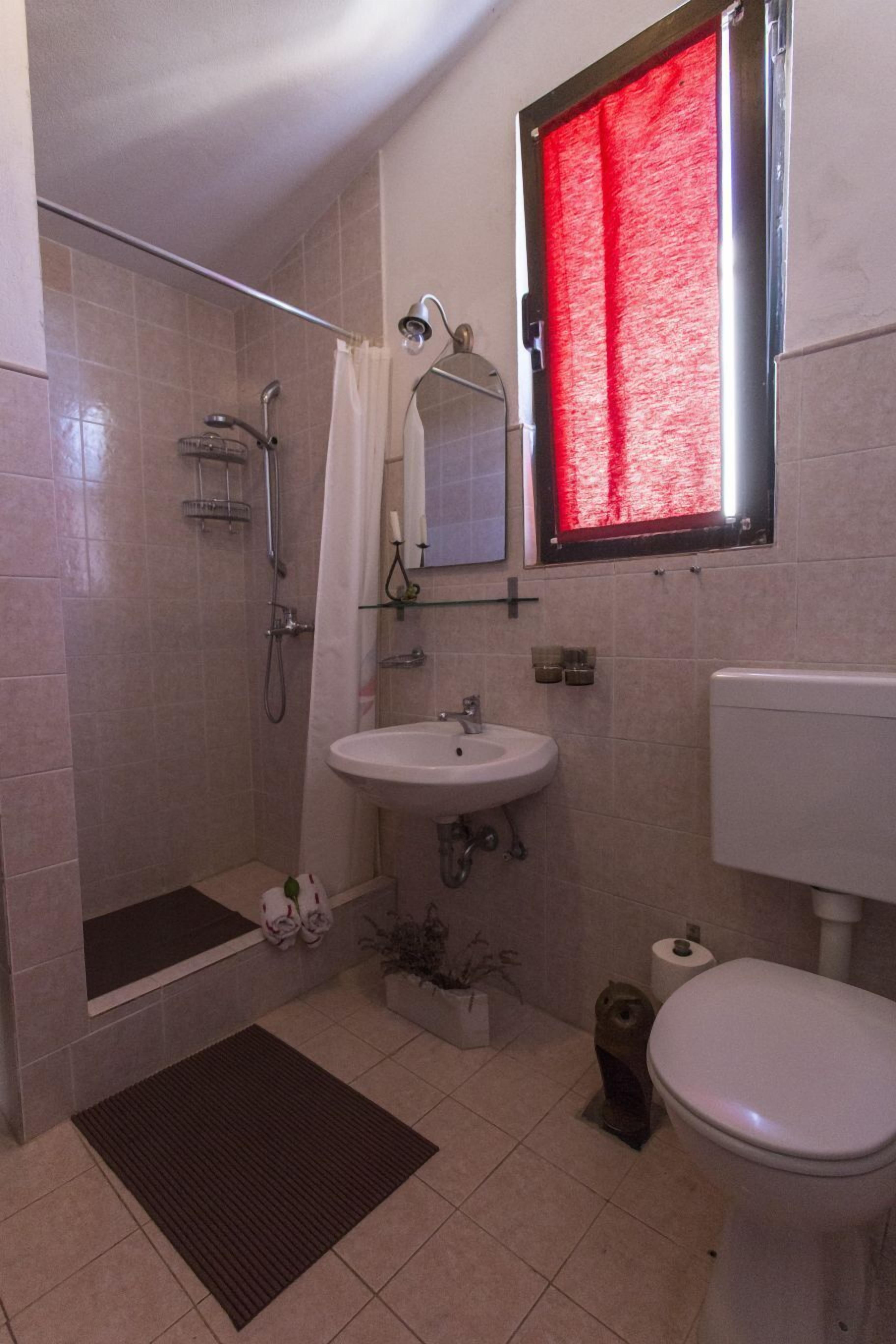 A bathroom with a shower, sink, toilet, and a window with a red curtain.