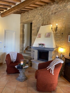 A cozy living room with a stone fireplace, two red armchairs, a glass coffee table with a candle, and a staircase leading to another room.
