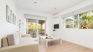 Ein modernes Wohnzimmer mit einer Couch, einem Couchtisch und einem Fernseher. Ein Balkon bietet Blick auf Bäume und eine Zimmerpalme.