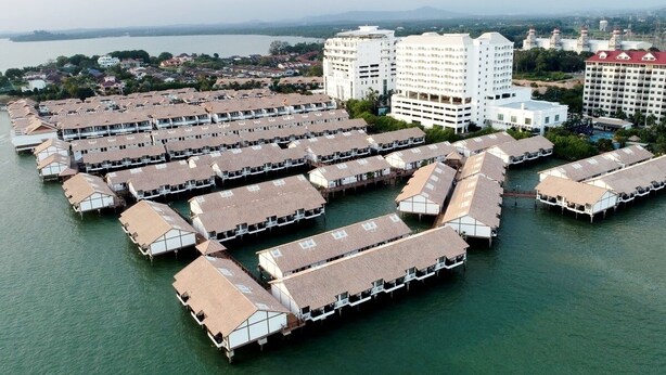 Vista aérea de un hotel frente al mar con varias cabañas sobre el agua dispuestas en filas.