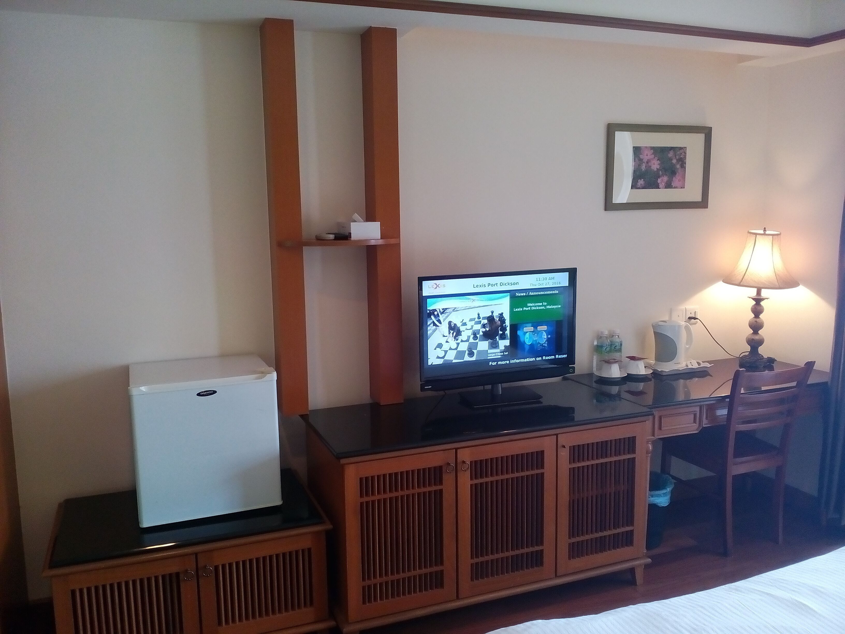 A hotel room with a bed, a desk with a computer, a refrigerator, a lamp, and a small shelf with items.