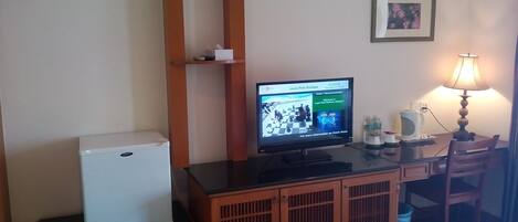 A hotel room with a bed, a desk with a computer, a refrigerator, a lamp, and a small shelf with items.