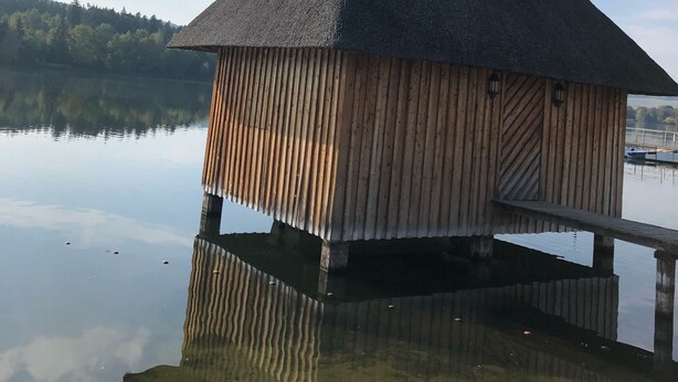 A wooden structure with a thatched roof on stilts over calm water, surrounded by a forested landscape.